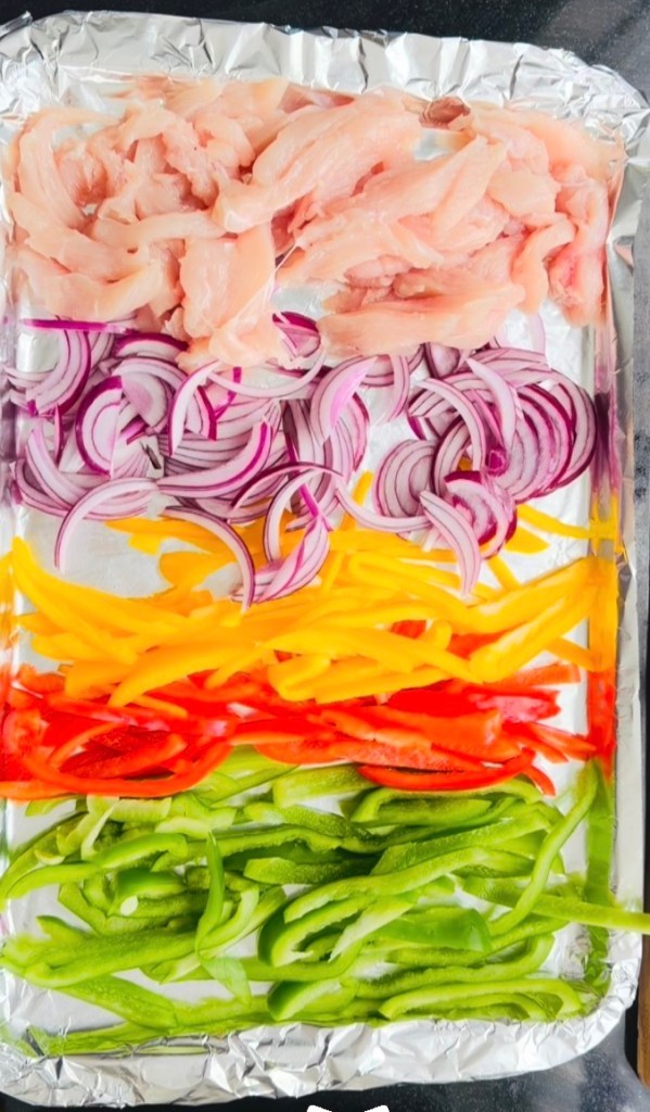 Raw julienne cut chicken breast, red onion and yellow, red and green capsicums arranged in rows on a foil-lined baking tray