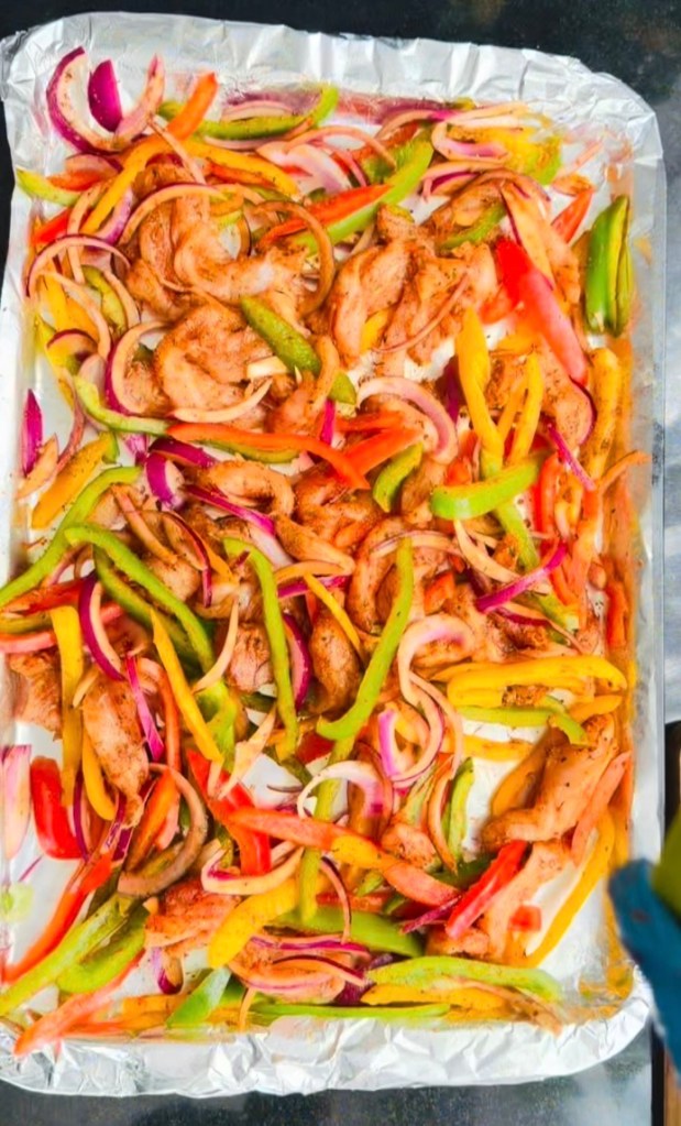Chicken strips, red onion and capsicums mixed with fajita seasoning on a foil-lined tray before baking
