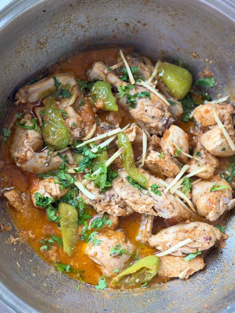 Chicken karahi in a steel karahi pan, topped with fried green chillies, slivers of fresh ginger and chopped coriander